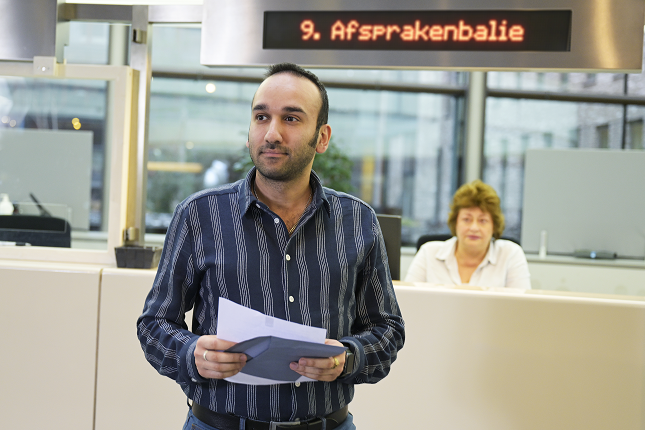 Een man met een envelop en een brief in zijn handen met op de achtergrond de balie van de Belastingdienst, met een bord waarop staat: afsprakenbalie.